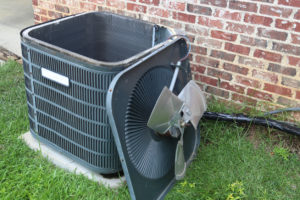 AC unit on the outside of a brick house. It is near the wall. The top part with the fan is removed, leaning against the unit. 